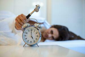 joven mujer intentos a descanso el alarma reloj con martillo, destruir el reloj. niña acostado en cama torneado apagado un alarma reloj con martillo en el Mañana a 5 de la mañana. foto