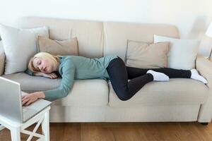 Exhausted young woman lying on sofa, using laptop, too tired or bored of online work at home, free space. Workaholism, chronic fatigue, overworking on remote job concept photo