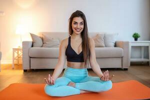 joven y atractiva mujer sonriente practicando yoga, sentada en medio ejercicio de loto, pose de ardha padmasana, haciendo ejercicio, usando ropa deportiva, sesión de meditación, interior completo, interior de casa foto