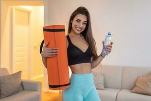 Beautiful young woman in sports wear is holding a yoga mat and a bottle of water, looking at camera and smiling photo