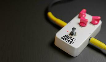 White Guitar Pedal, Overdrive, Pink Kinobs Connected by Two Cables on the Floor, against Black Background photo