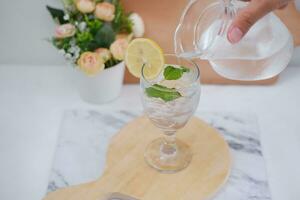 Pouring water into glass with mint and lemon on white marble table photo