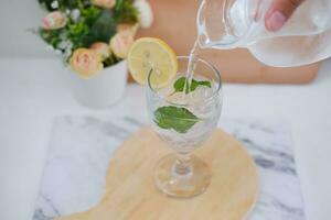 Pouring water into glass with mint and lemon on white marble table photo