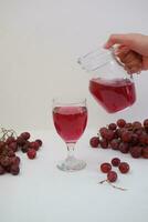 Red grape juice pouring into a glass with fresh grapes on white background photo