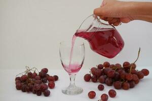 Red grape juice pouring into a glass with fresh grapes on white background photo