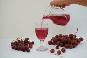 Red grape juice pouring into a glass with fresh grapes on white background photo