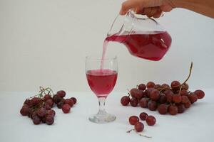Red grape juice pouring into a glass with fresh grapes on white background photo