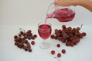 Red grape juice pouring into a glass with fresh grapes on white background photo