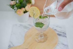 Pouring water into glass with mint and lemon on white marble table photo