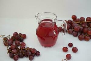 uva jugo en un vaso jarra con Fresco uvas en un blanco antecedentes foto