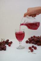 Red grape juice pouring into a glass with fresh grapes on white background photo