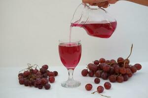 Red grape juice pouring into a glass with fresh grapes on white background photo