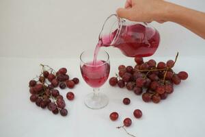 Red grape juice pouring into a glass with fresh grapes on white background photo