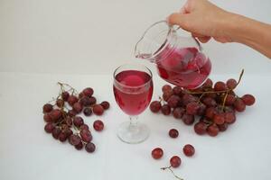Red grape juice pouring into a glass with fresh grapes on white background photo