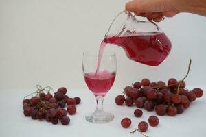 Red grape juice pouring into a glass with fresh grapes on white background photo