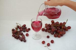 Red grape juice pouring into a glass with fresh grapes on white background photo