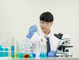 retrato asiático hombre estudiante científico vistiendo un médico vestido en el laboratorio mirando mano a químico. causado por mezcla reactivos en científico investigación laboratorios con prueba tubos y microscopio en el mesa foto