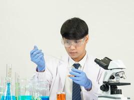 retrato asiático hombre estudiante científico vistiendo un médico vestido en el laboratorio mirando mano a químico. causado por mezcla reactivos en científico investigación laboratorios con prueba tubos y microscopio en el mesa foto