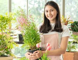 Portrait garden beautiful pretty young Asian woman wearing white blouse with long black hair and smile fresh happy relax bright smile look hand holding pot small tree leafgreen plant in room shop photo