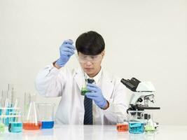 retrato asiático hombre estudiante científico vistiendo un médico vestido en el laboratorio mirando mano a químico. causado por mezcla reactivos en científico investigación laboratorios con prueba tubos y microscopio en el mesa foto