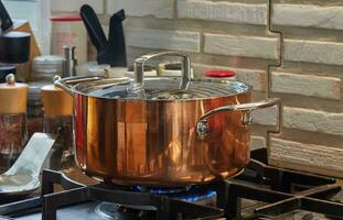 Copper saucepan with lid with dish is cooked over fire on gas stove. Steam rises above the pot photo