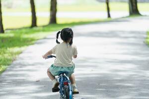 vista lateral trasera de una niña asiática aprendiendo a andar en bicicleta en el jardín del parque. concepto de educación para niños que practican ciclismo en el parque, concepto de deporte para bebés. foto