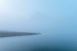 Traveler man standing alone in blue fog with rocky mountains by the lake in the morning photo