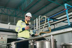 caucásico técnico ingeniero hombre en uniforme con tableta comprobación y controlar caldera tanques y líquido tubería en producción línea a fábrica foto