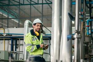 Caucasian technician engineer man in uniform with tablet checking and control boiler tanks and liquid pipeline in production line at factory photo