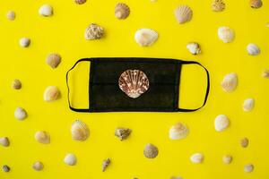 Black medical face mask and seashells on a yellow background. Flat lying, copy space, holiday cancellation, pandemic, tourism. photo