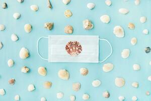 Black medical face mask and seashells on blue background. Flat lying, copy space, holiday cancellation, pandemic, tourism. photo