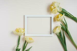 White wooden photo frame framed with yellow tulip flowers on a light table. Flat lay, card