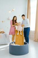 joven hermosa caucásico familia jugando con su un año de edad hija en el para niños habitación. el niña diapositivas abajo el diapositiva dentro el piscina con pelotas foto