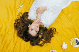 Top view of a beautiful romantic woman in a dress, lying on the green grass on a yellow blanket. Picnic in the park. Fruit on a plate. Summer holidays. photo