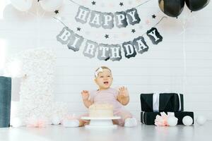 un uno año antiguo niña se sienta en el piso cerca un dulce pastel en honor de su primero cumpleaños, intentos el pastel y sonrisas, se ríe foto