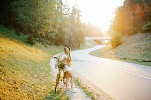 retrato de un contento hermosa joven niña con un bicicleta y flores en el antecedentes de un bosque por el la carretera en el luz de sol en el abierto aire foto