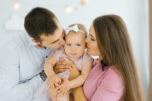 joven contento padres sostener su un año de edad hija en su brazos y Beso su en el mejilla foto