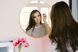 Girl with dark hair stands in front of a mirror and does makeup, holding a brush in her hand, creating an image, beauty and style photo