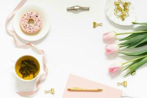 elegante plano laico para un mujer Moda blogger lugar de trabajo de un persona de libre dedicación o oficina obrero, un cubierto de rosa diario, un bolígrafo, un taza de verde té, un rosado rosquilla, un ramo de flores de rosado primavera tulipanes foto