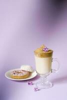 Beautiful dalgona drink a frothy coffee in a transparent mug and flowers of lilac on a purple background. Sweets on a plate, Donat and tender marshmallows. Copy space photo