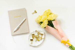 Trending minimalistic flat lay of a blogger's workplace. Notepad, pen, gold-colored paper clips and a bunch of yellow tulips on a white background. Top view photo