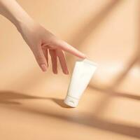 Woman's hand touches a mockup of white tube of cream. Unlabeled packaging for cosmetics in sunlight on beige background. Concept of skincare. Harsh shadows photo