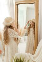 A beautiful teenage girl with long hair measures a straw hat in front of a mirror. Self-admiration of a blonde. photo