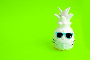 Pineapple in a medical mask and sunglasses on green neon background. Summer in Covid-19. Vision for the summer of 2020 during the coronavirus pandemic. photo