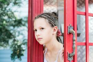 un triste pequeño niña con trenzas afro mira fuera desde un antiguo Inglés teléfono puesto. generacional contraste foto