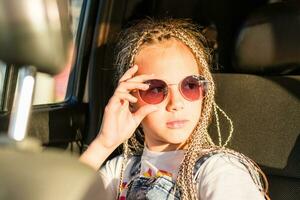 grave adolescente niña con trenzas afro sostiene Gafas de sol con su mano y mira fuera el ventana mientras sentado en el coche. la carretera viaje foto