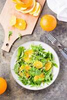 Diet vegetarian vitamin salad of orange slices and a mix of arugula, chard and mizun leaves on a plate and a cutting board with peeled orange on the table. Top and vertical view photo