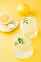 Sparkling water with lemon, melissa and ice in glasses and lemon slices on a saucer on a yellow background. Alcoholic drink hard seltzer. Vertical view photo