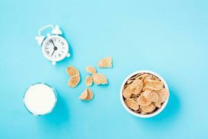 un cuenco de cereal, un vaso de Leche y un alarma reloj en un azul antecedentes. programado desayuno. parte superior ver foto