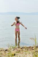 Adolescente niña en un traje de baño abanico sí misma con un Escoba de césped imitando bañera procedimientos en el soleado río banco en verano foto